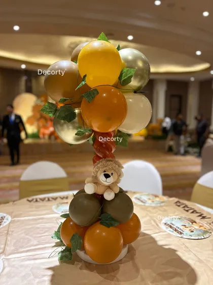 A detailed look at a table centerpiece. It features a balloon arrangement in the theme's color palette of orange, gold, and brown, topped with a cute lion toy.