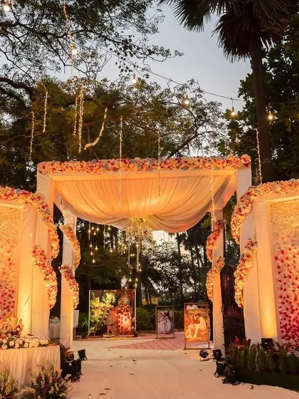 A grand entrance for a garden wedding, featuring large, illuminated panels covered in flowers. The warm lighting and string light canopy create a magical and impressive first look.