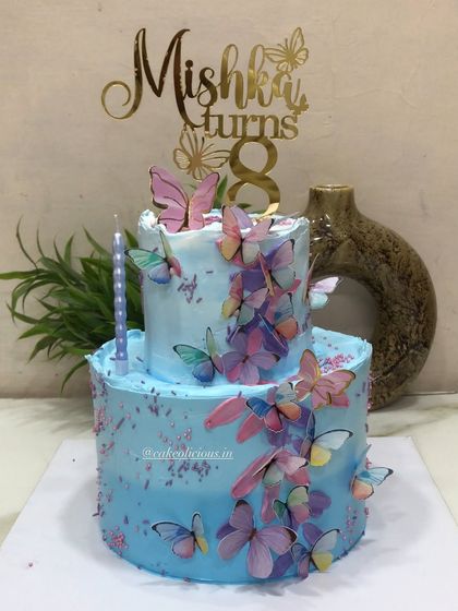 A pretty two-tier butterfly-themed cake for an 8th birthday. This 3kg chocolate mousse cake is covered in blue whipped cream and a cascade of colorful, edible butterflies, complete with a custom name topper.