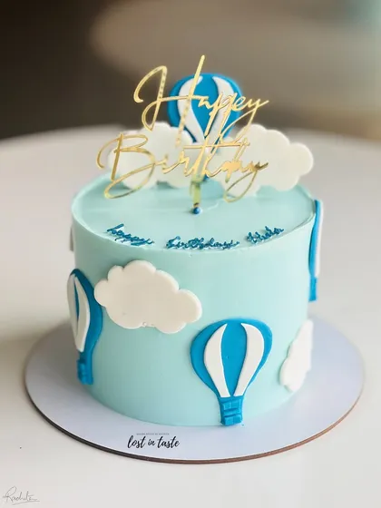 A whimsical hot air balloon themed cake. The light blue buttercream sky, fluffy clouds, and a golden "Happy Birthday" topper make it a delightful choice for a child's celebration.