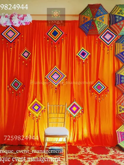 A colorful Mehndi ceremony backdrop. The bright orange drape is decorated with traditional Rajasthani-style hangings and colorful umbrellas, creating a festive and vibrant photo corner.