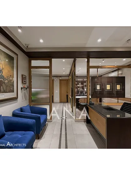 The reception and waiting area of the Jain Cosphere Office. The design uses a palette of beige, wood, and dark green, with blue sofas adding a pop of color. The space is designed to feel open yet private.