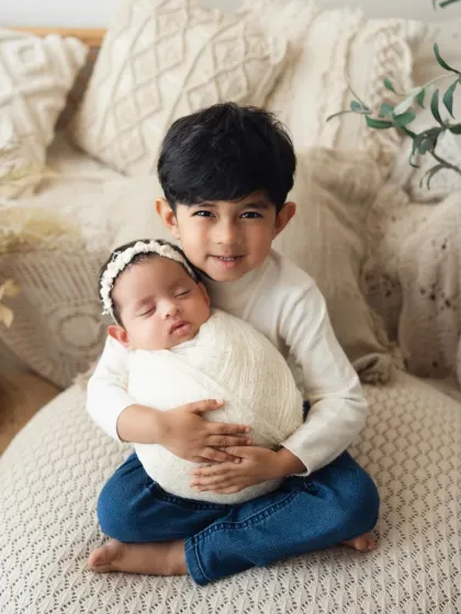 An older brother holds his newborn sibling carefully on a soft, boho-inspired bed setup. This is a beautiful example of a newborn and older sibling pose.