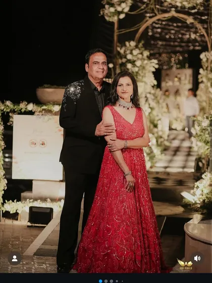A couple dressed for a wedding reception in our coordinated designs. The gentleman wears a custom black suit with unique shoulder embellishments, complementing his partner's elegant red gown.