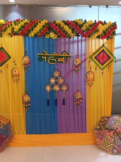 A colorful Mehandi backdrop with yellow, blue, and purple drapes. The setup is decorated with traditional hangings and Rajasthani umbrellas, perfect for a lively event.