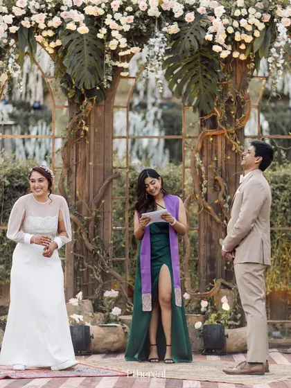 A friend officiates the wedding, reading from a script as the bride and groom look on with smiles. This highlights the trend of personal, friend-officiated ceremonies that are deeply meaningful.