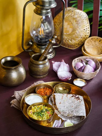 A traditional meal of Jhunka Bhakar served on a brass plate, with an antique lantern in the background. The styling evokes a sense of nostalgia and the timelessness of Maharashtrian village life.