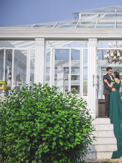 A wide shot showing the couple on the steps of an elegant glasshouse, framed by lush green bushes. This image captures the beauty of the location and the couple's grand entrance.