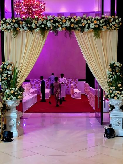 An elegant entrance featuring soft cream drapes and classic white urns filled with vintage-style florals. The design creates a warm and sophisticated welcome for a wedding at the Taj Santacruz.