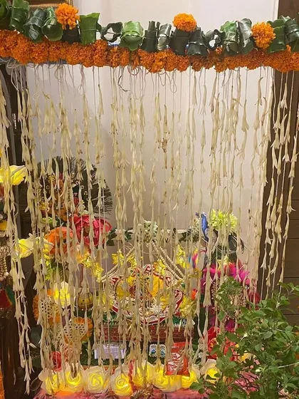 A small mandap created for a home pooja. The frame is wrapped in marigold garlands, and strings of rajnigandha (tuberose) hang in the background for a traditional look and fragrance.