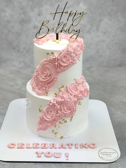 A two-tiered cake with a beautiful cascading floral design. The pink piped roses flow diagonally across the cake, accented with gold leaf for a touch of elegance.