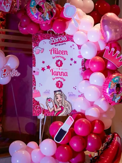 A personalized welcome board for Aileen and Tanna's Barbie-themed first birthday party. The design is surrounded by pink balloons and themed cutouts like lipstick and a crown.