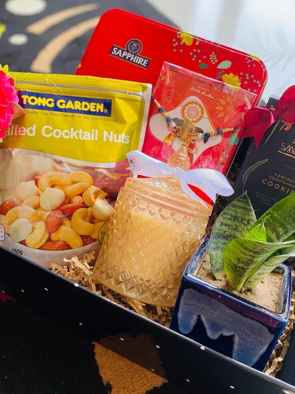 A thoughtful and diverse gift box combining treats, wellness, and a touch of green. This hamper includes salted cocktail nuts, cookies, a scented candle, a traditional rakhi, and a small snake plant for a lasting keepsake.
