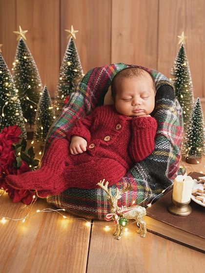 A festive Christmas-themed newborn shoot, with the baby dressed in a cozy red outfit, surrounded by miniature trees, fairy lights, and holiday props.
