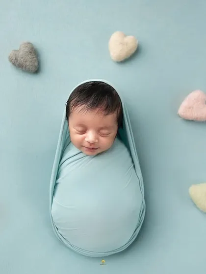A minimalist portrait of a swaddled baby on a simple blue background, surrounded by pastel-colored hearts. The focus is entirely on the baby's sweet, sleeping face.