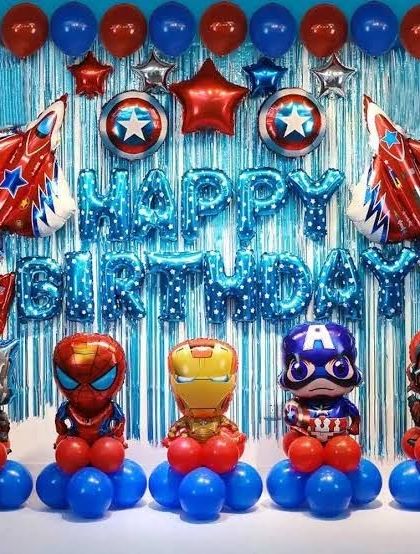 An action-packed Avengers birthday decoration. This setup includes a blue tinsel curtain backdrop, "Happy Birthday" foil letters, and balloon stands featuring the full team of superheroes like Iron Man, Captain America, and Spider-Man.