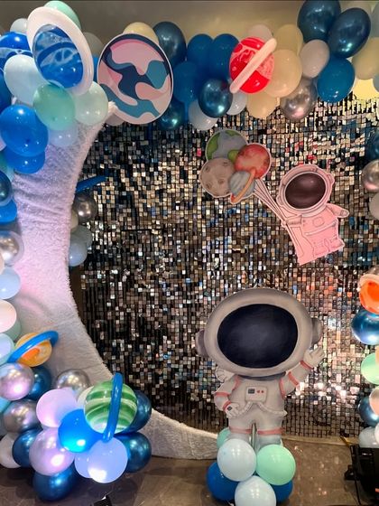 A fun photo-op area with an astronaut cutout where guests can place their face for a picture, set against a silver shimmer wall and planet balloons.
