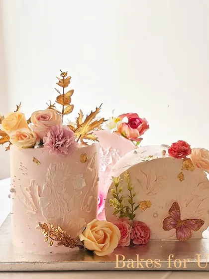 A unique two-part cake design in a soft pink, decorated with stenciling, fresh flowers, and gold accents. This modern arrangement is perfect for a stylish celebration.