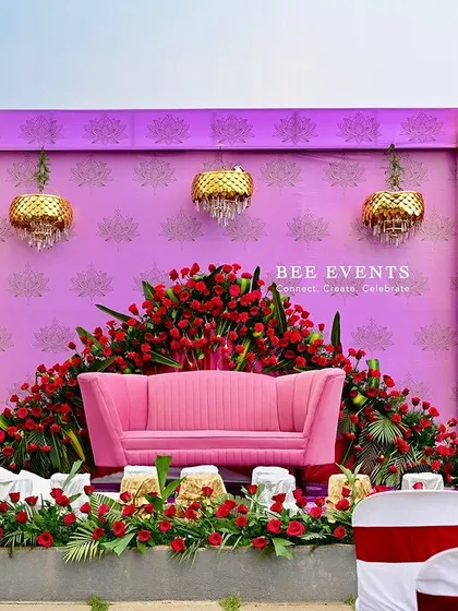 A vibrant and regal stage setup perfect for a reception or engagement. The design uses a bold color palette of pink and red, with a plush pink sofa, abundant red roses, and elegant golden chandeliers for a touch of royalty.