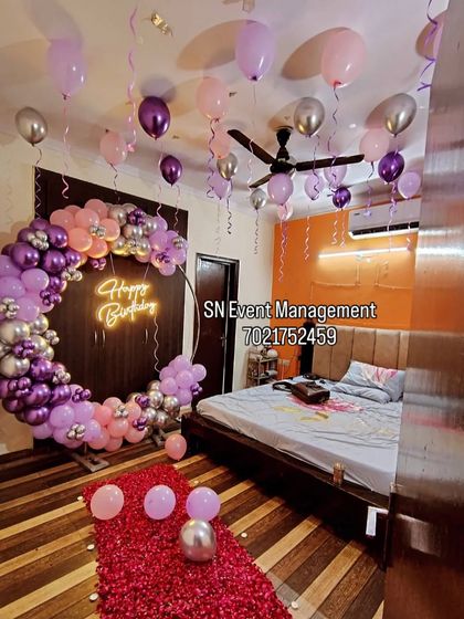 An elaborate and romantic birthday room decoration. This setup includes a full balloon ring in purple and pink, balloons on the ceiling, and a path of rose petals leading to the bed.