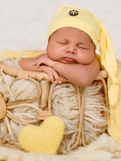 A newborn with chubby cheeks and a cute sleepy hat rests their head on their hands in a basket.
