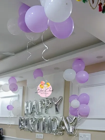 A simple birthday setup with silver 'Happy Birthday' foil letters and bunches of purple and white balloons on the ceiling.