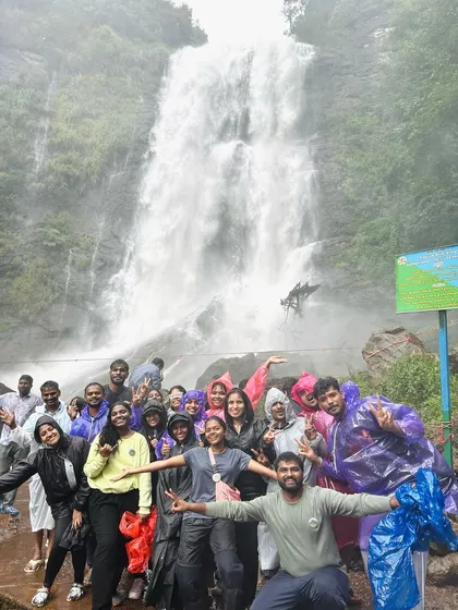 A joyful group in raincoats posing with arms outstretched in front of a massive waterfall during a monsoon trip. This is the pure happiness of a waterfall visit.