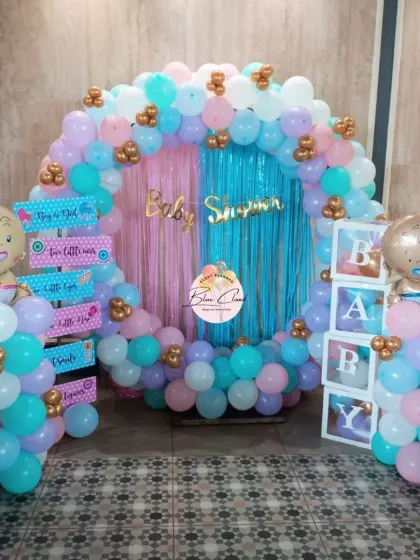 A beautiful gender reveal party setup with a full balloon ring in pastel pink, blue, and purple. The decor includes 'BABY' blocks, themed foil balloons, and a shimmer curtain backdrop.
