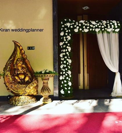 A simple and elegant entrance decoration for an engagement ceremony. A Ganesha idol is placed alongside traditional props and a beautiful floral arch with a white curtain.
