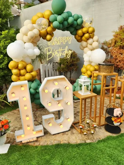 A rustic and cheerful 19th birthday setup in the garden. This design combines a green and gold balloon arch with large marquee numbers and wooden decor elements for a natural and celebratory feel.