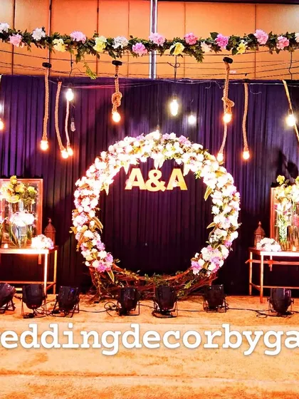 This is the stage for a Sangeet night, featuring a large floral wreath with the couple's initials. The backdrop of dark drapes is accented with hanging Edison bulbs for a rustic yet modern feel.