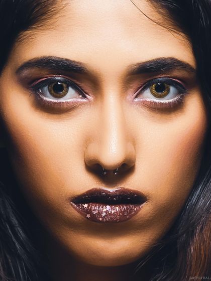 An intense, direct-to-camera beauty portrait. The focus is on the model's striking eyes and the creative, sparkling lip makeup.