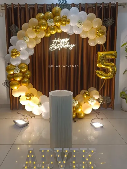 A simple yet elegant 5th birthday setup. The golden hoop is decorated with a white and gold balloon arch, with a 'Happy Birthday' neon sign and marquee letters creating a warm glow.