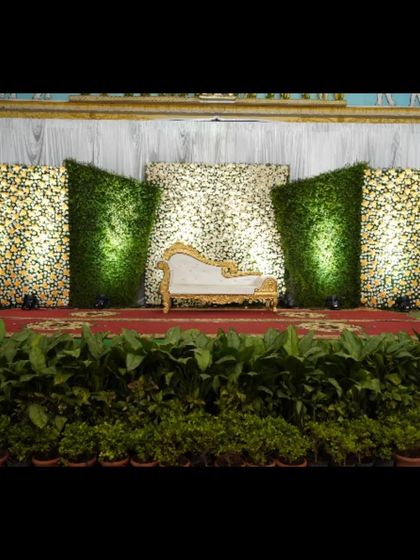This unique stage design features panels of greenery and flowers with dramatic angles and uplighting. The central flower wall provides a textured backdrop for the ornate white and gold seating.
