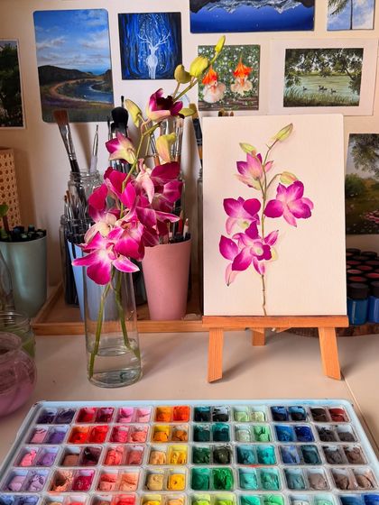 The finished pink orchid painting, displayed on a mini easel in my studio. It sits beside the real flower and my palette, showing the full story from inspiration to final artwork.