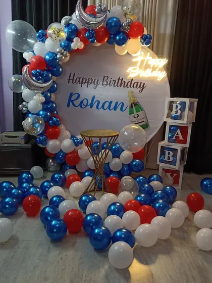 A personalized birthday setup for Rohan, featuring a custom printed circular backdrop. The decor is completed with a red, white, and blue balloon arch, 'BABY' blocks, and a 'Happy Birthday' neon sign.