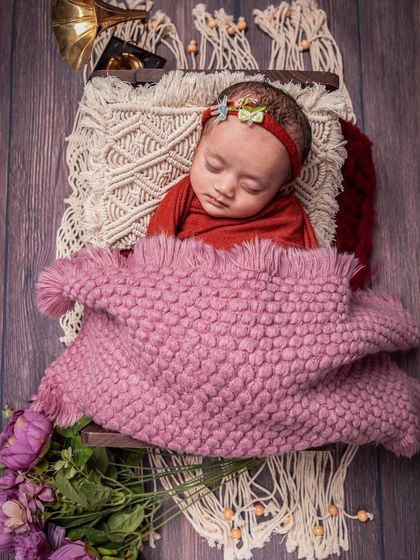A cozy, rustic portrait of a newborn sleeping on a wooden crate with a macrame runner. The rich red wrap and soft pink blanket add warmth and color to this charming setup.