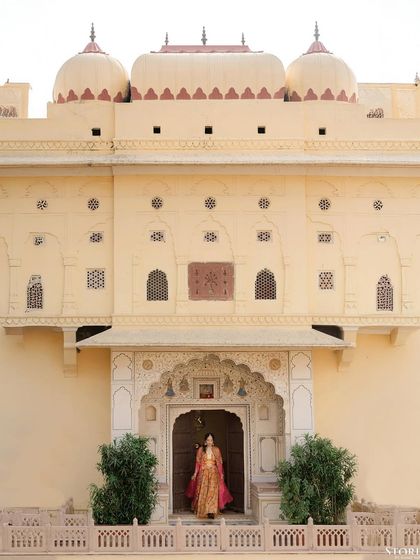 A wide shot of the bride, Aashi, standing alone in the grand entrance of a palace. This portrait emphasizes her regal presence and the magnificent scale of the venue.