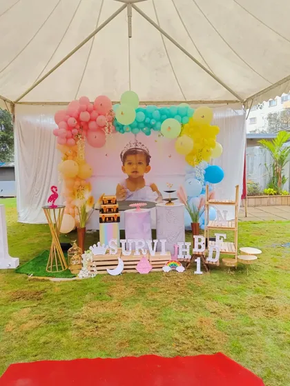 An outdoor party setup under a canopy for Survi's first birthday. The decoration features a pastel rainbow balloon arch and a large photo of the birthday girl.