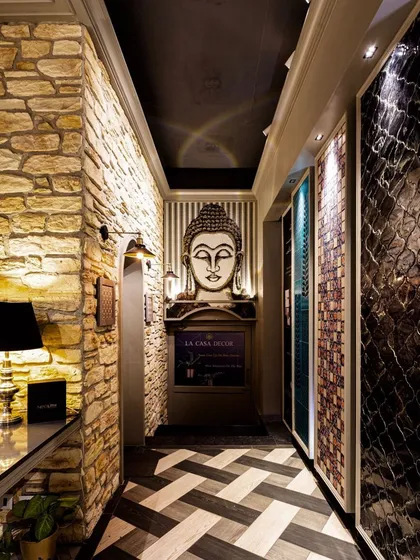 The entrance corridor of the La Casa tile studio, where lighting creates a serene and welcoming path. A combination of a classic wall sconce, a soft table lamp, and focused spotlights on the Buddha statue and tile panels creates a layered and inviting entryway.