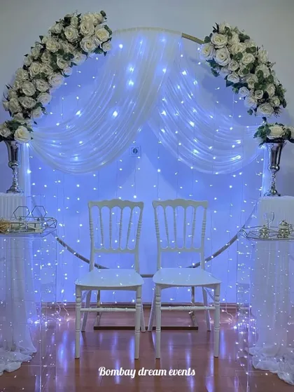An elegant anniversary decor setup featuring a circular frame with white drapes, fairy lights, and white roses, perfect for an intimate celebration.