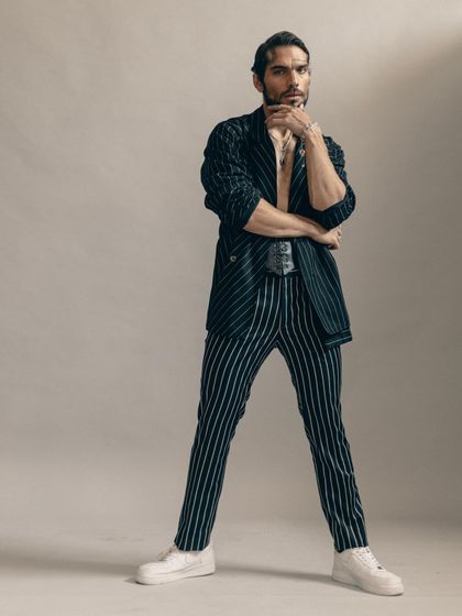 A full-length standing pose in the pinstripe suit, styled with a corset for a high-fashion edge. The look combines classic menswear with avant-garde elements for a memorable editorial image.
