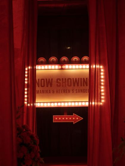 The "Now Showing" marquee sign, seen through parted red velvet curtains, creating a dramatic and inviting entrance for the Sangeet night.