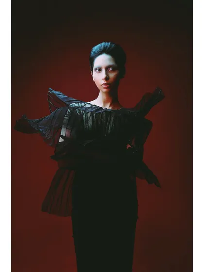 A direct, confrontational portrait of 'Athena'. The model, styled in a dramatic black pleated top, looks straight at the camera. The red lighting adds an intense, emotional undercurrent.