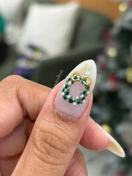 A close-up of a single nail featuring a hand-crafted Christmas wreath made from tiny green and white beads with a 3D gold bow.