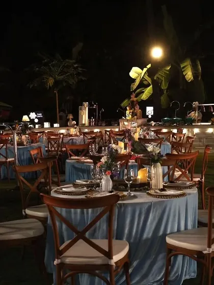 An evening view of the setup for Ezekiel's First Holy Communion. The outdoor lawn was set with tables draped in blue linen, wooden cross-back chairs, and a well-lit buffet counter in the background.