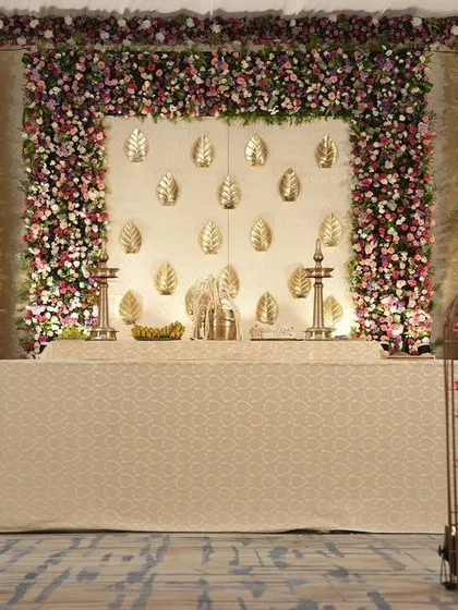 An elegant indoor mandap for a South Indian wedding, featuring a backdrop of golden leaves, flanked by dense walls of pink and white flowers and lit by hanging geometric lamps.