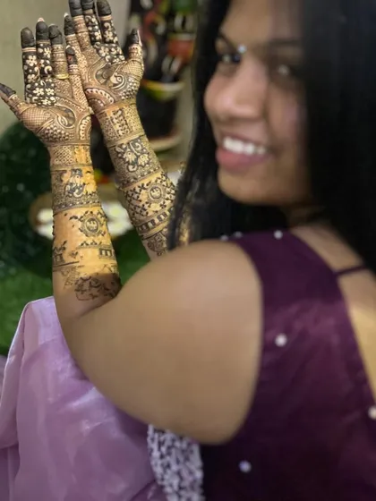 A smiling bride showing her full bridal mehendi. The design is traditional, with detailed patterns covering her hands and forearms.