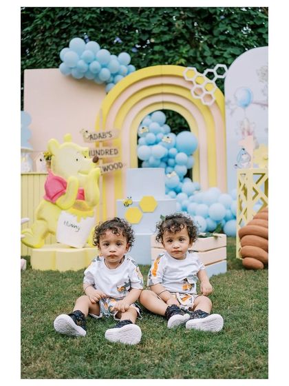 The adorable birthday twins sitting in front of their Winnie the Pooh party setup. The backdrop features a pastel rainbow, a Pooh bear prop, and a sign for 'Adab's Hundred Acrewood'.