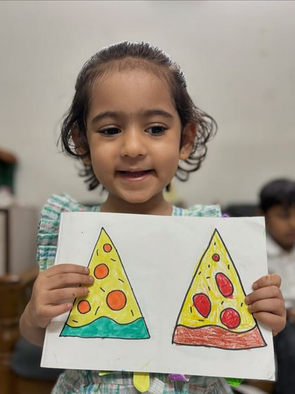 Learning shapes can be delicious. A junior KG student happily shows her drawing of pizza slices, a fun exercise we do to practice triangles and circles while learning to colour within the lines.